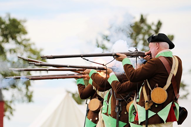 Men dressed in brown jackets with green insides and black hats fire muskets as white smoke and sparks billow out of them