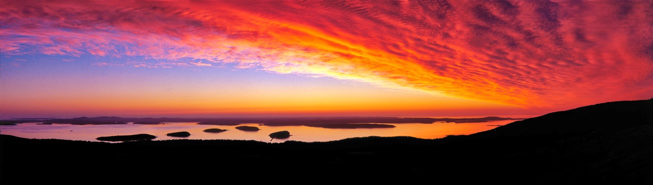 Panorama of sunrise with bright yellow, red, and orange clouds filling the sky at right, a water inlet with islands at center, and the silhouette of a mountain ridge framing the foreground
