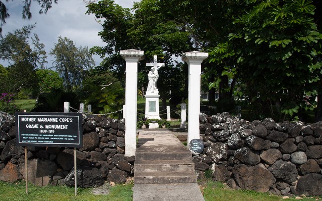 White stone grave marker with saint holding Jesus as he hangs from cross. Historic marker reads "Mother Marianne Cope's Grave & Monument (1838-1918)"