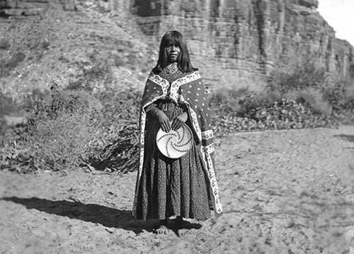 In a black and white photo, a woman stands barefoot on the sand wearing historical Havasupai clothing.