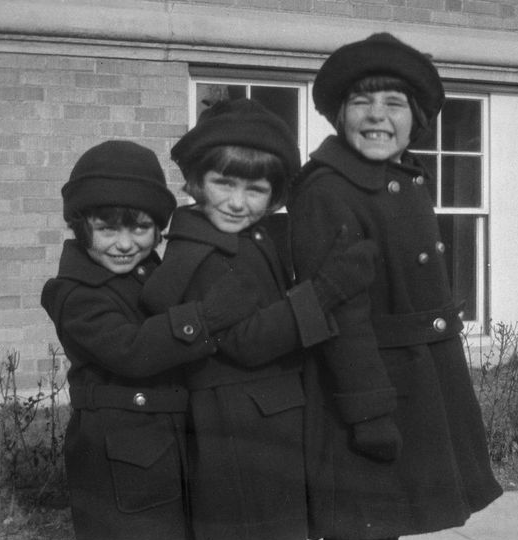 Eunice, Kick, and Rosemary, ca. 1925 A black and white photo of three smiling girls in matching dark coats, hats, and bob haircuts standing in order of height, in front of a brick building. The two smaller girls each hold the arm of the girl in front. The tallest girl grins widely.