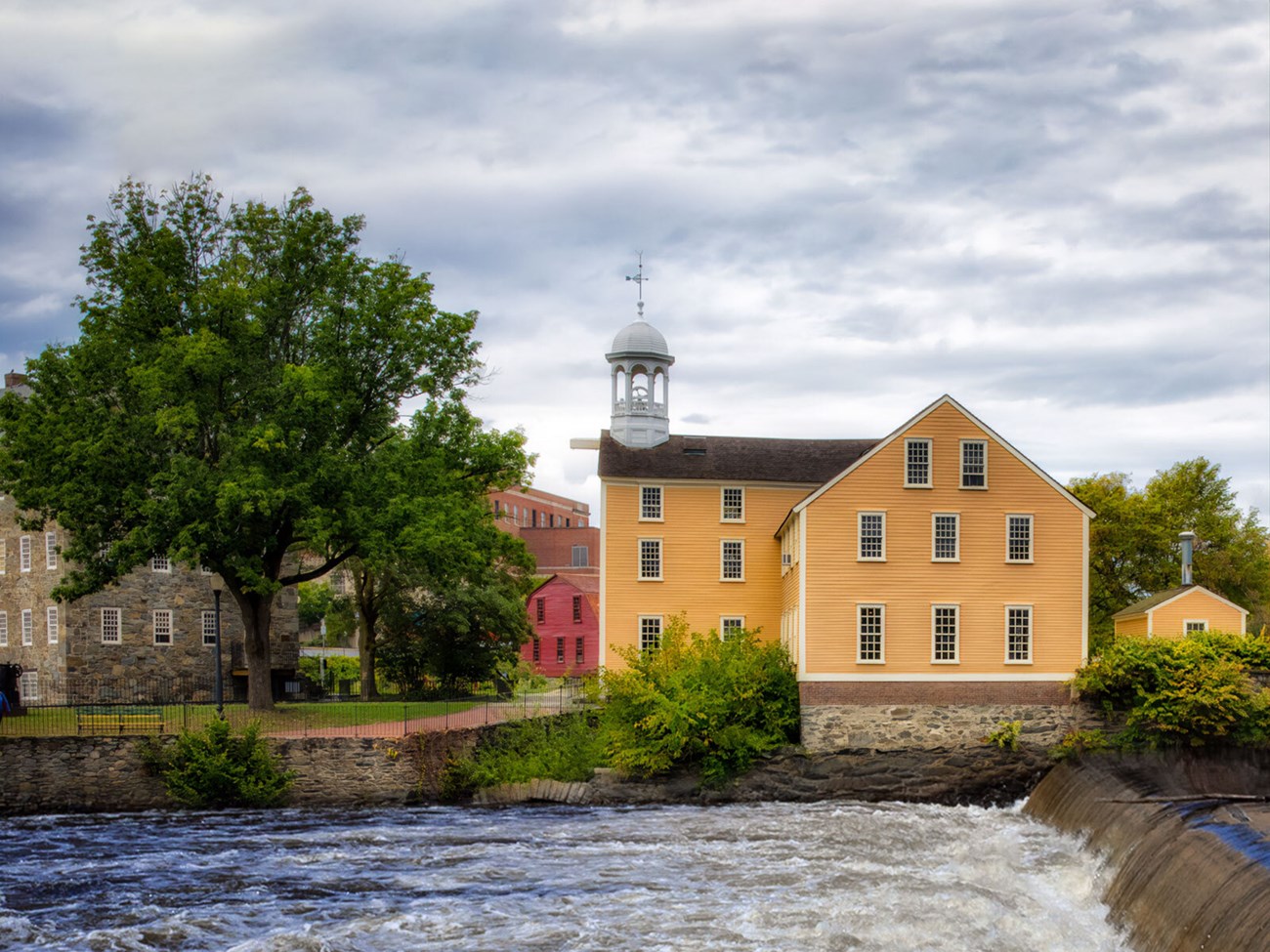 Slater and Wilkinson Mills Cloudy day view looking across the river at a yellow three-story building (center right). On the left is a multi-story stone buildingpartly obscured by a large tree, and in the back center, there's a bright red two-story house.