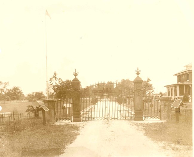 Early 1900s image of ornate gates and entry road to Chalmette National Cemetery, metal signs, flagpole, and brick two-story lodge.