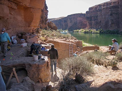People sit and stand around an archaeological site next to the river.