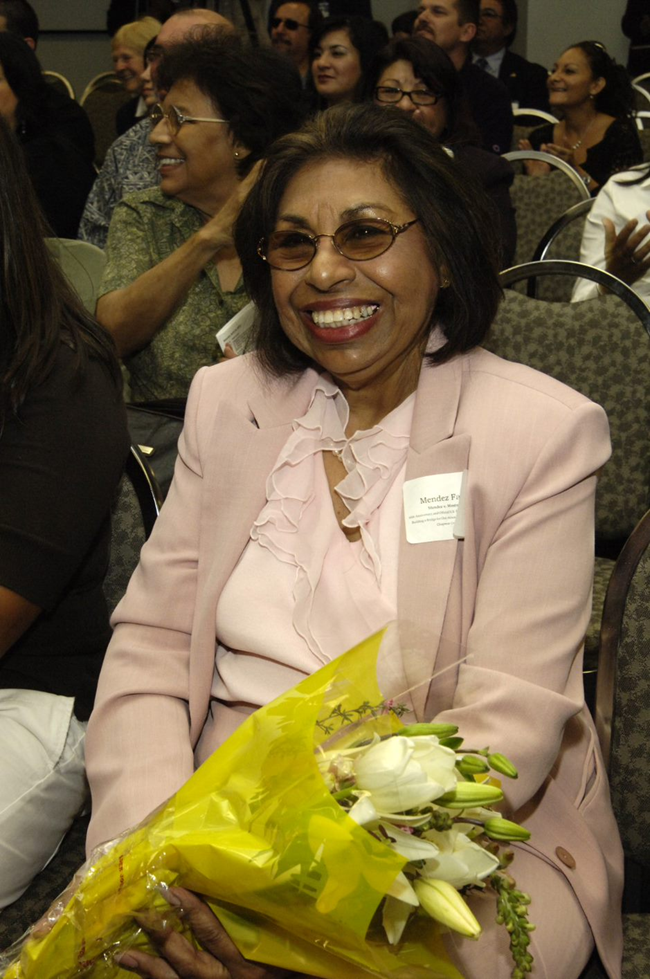 Sylvia Mendez celebrates the release of the US Postal Service’s Mendez v. Westminster commemorative stamp at Chapman University, 2007. Courtesy of Chapman University, Frank Mt. Pleasant Library of Special Collections and Archives. Color photo of Sylvia Mendez as an adult wearing a pale pink skirt suit and blouse and lightly tinted oval glasses. She is seated with a bouquet of flowers and smiles toward the camera.