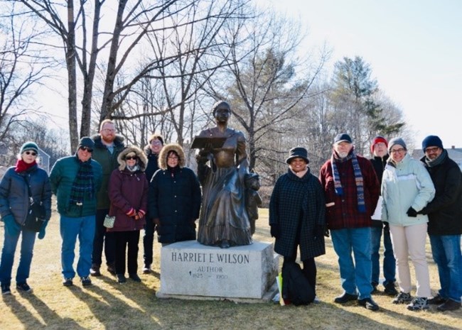Bronze memorial statue, with six people standing to its left and five to its right, all in winter garb. Bare trees in background with bright sky shining behind. Stone base of memorial is engraved with text: "Harriet E. Wilson, Author"