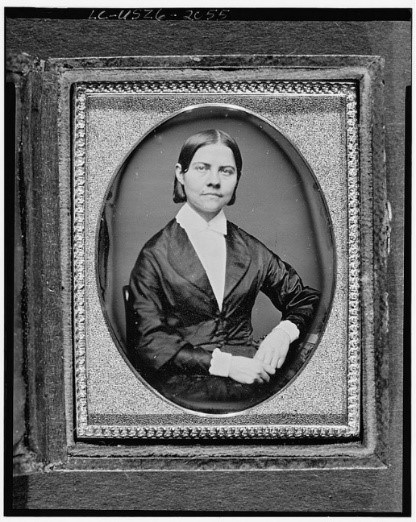 Black-and-white portrait photograph of seated woman in oval vignette-shaped matting and frame. She wears aa dark collared robe overcoat or dress over collared white shirt, slick center-parted hair, and a matter-of-fact expression.