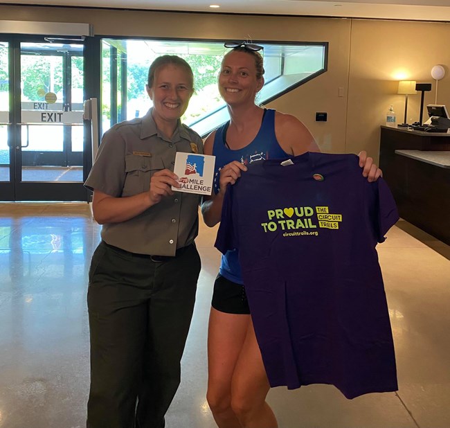 a uniformed park ranger and a woman in athletic clothes smile and hold a t-shirt and a magnet