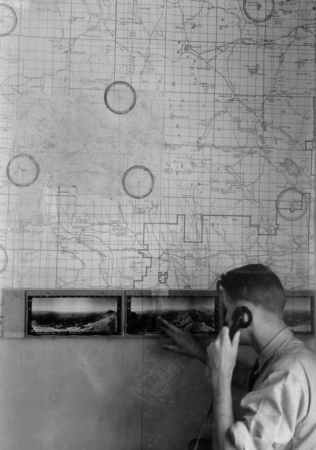 A man on the telephone points to panoramic photos below a large map on a wall.