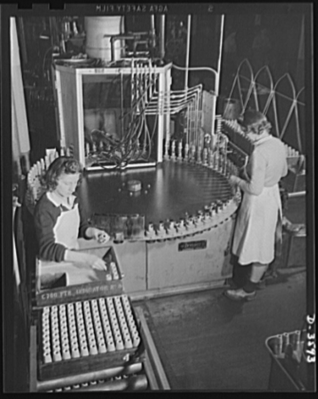 Two white women operate a machine that paints ammunition shells. Shells rotate along the outer edge of a circular track at the base of the machine to be painted. On the left, a woman adds completed shells to a tray. At right, the woman loads the track.