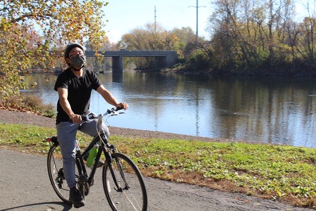 A person wearing a helmet and face mask stands stopped over bike looking forward on paved trail. Greenery and river to the person's right.