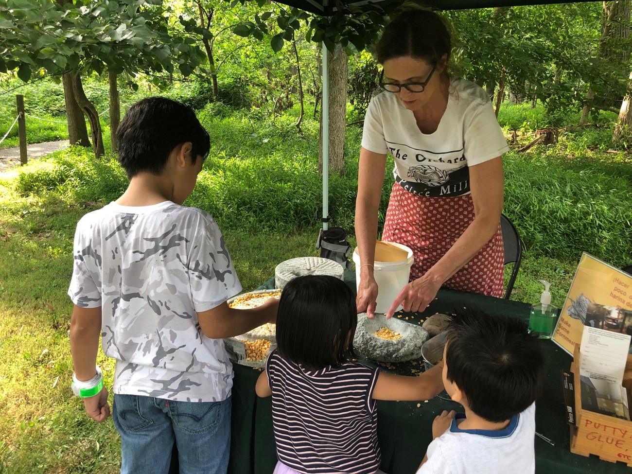 lt= A woman shows three children how corn is ground using a mortar and pestle