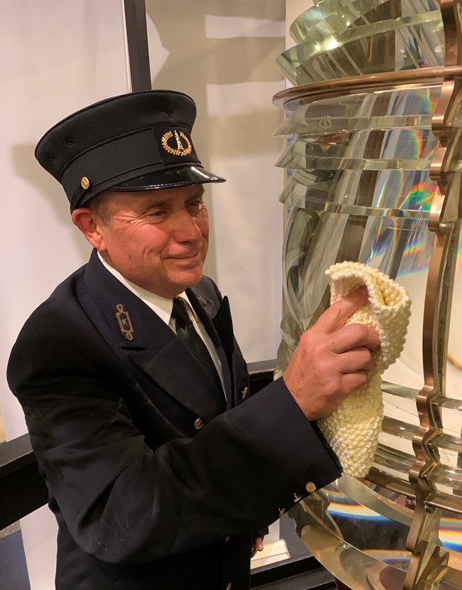 A man dressed in a black lighthouse keeper uniform and hat polishes a glass Fresnel lens.
