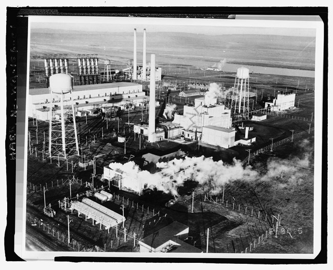 Black and white factory area. Multiple smokestacks with smoke rising and two water towers.