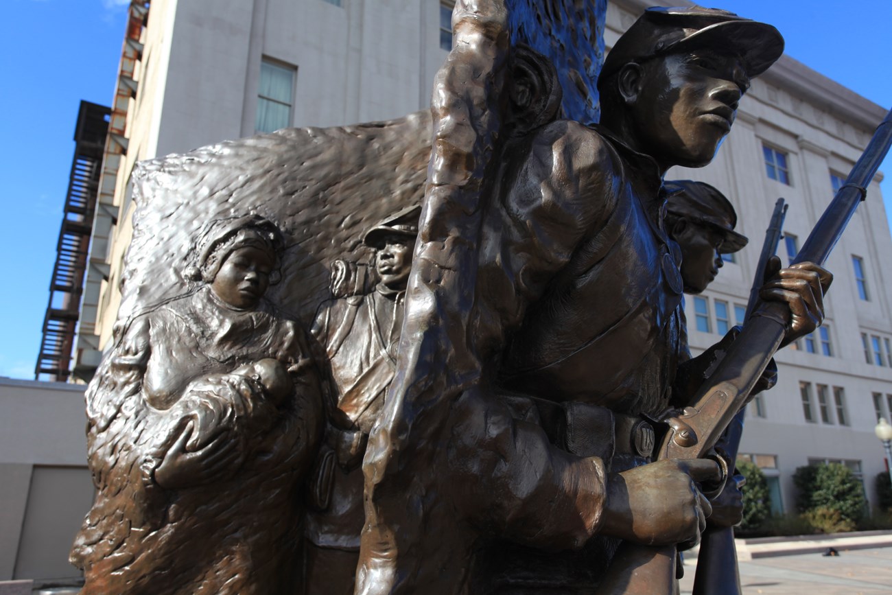Close-up of a statue showing Black soldiers in the Civil War on one side, protecting a woman holding a baby on the other.