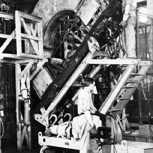 Two men in work uniforms wearing gloves inside a warehouse-type building. One man stands on the ground and the other on a raised platform a few feet above. The men are dislodging an Alpha calutron magnet.