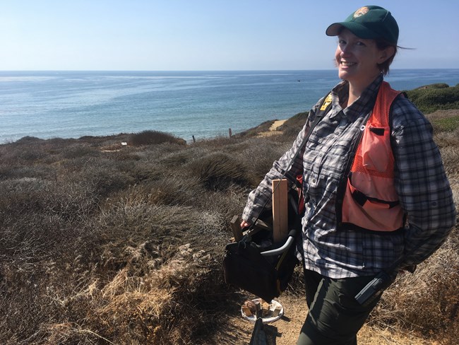 Woman in field clothes, orange safety vest, and green ballcap with the NPS arrowhead on it stands smiling in front of the green guide fencing of a herpetofauna pitfall array at Cabrillo National Monument. Blue ocean stretches to the horizon behind her.