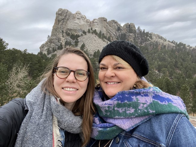 Amy and her mom stand smiling wearing scarves in front of Mount Rushmore National Memorial on a cold, gray day.