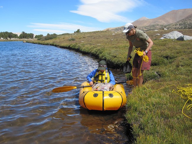 One woman wearing a life vest sits in a small raft at the edge of a lake while another woman stands on shore holding rope that is used to haul raft back to shore if any problems arise.