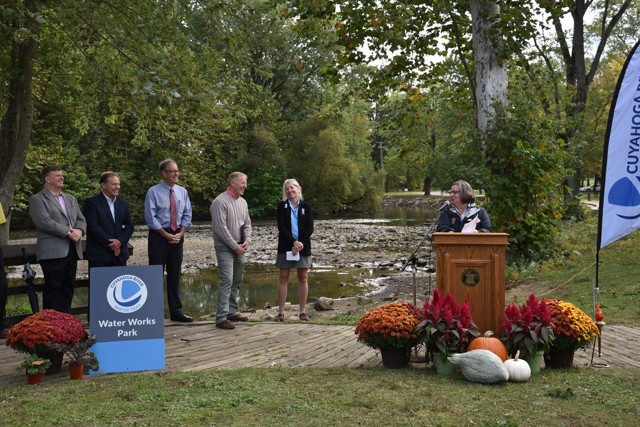 A woman stands behind a podium looking at distinguished individuals