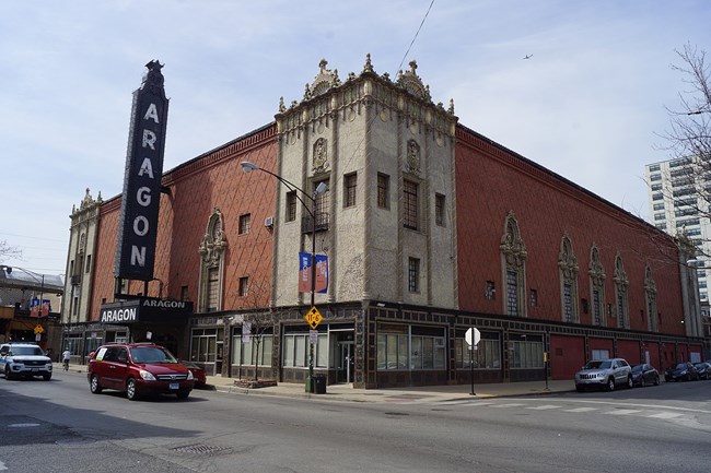 Corner view of the Aragon Ballroom. The building is clad with stucco in the Spanish Baroque style. Most of the exterior walls are red in color with ornate corner sections in a white. The venue's name is displayed vertically on a large sign at left.