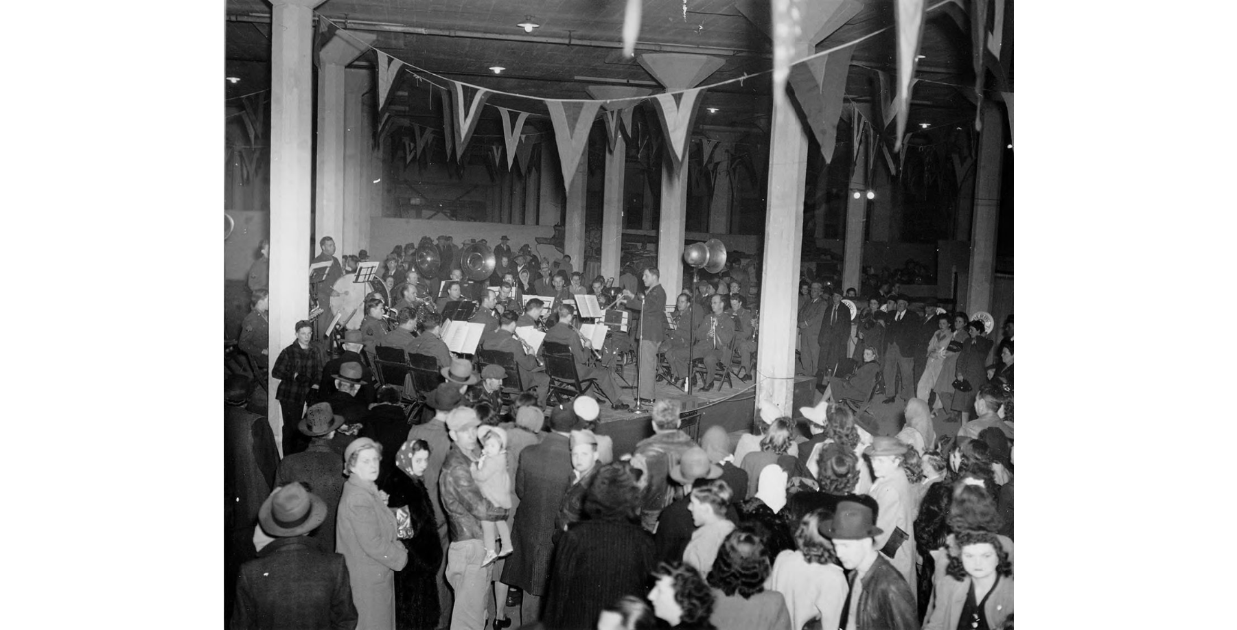 Black and white photo of a large room decorated with penants and packed with people. In the background is a band on a low stage.