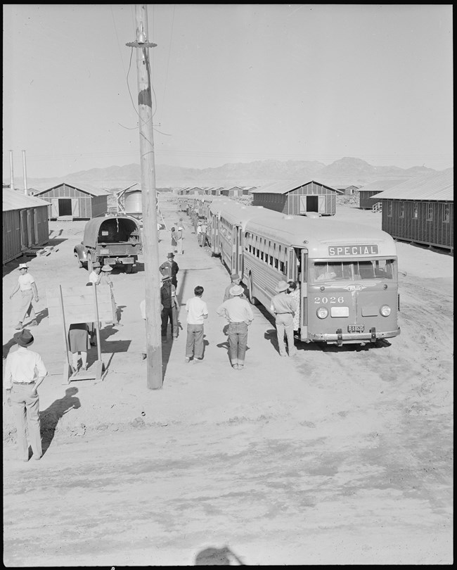 Japanese Americans disembark from a long row of buses that stretches off into the distance. The buses are parked in a desolate landscape between rows of low barrack buildings and an electrical pole.