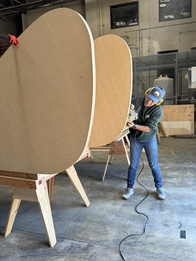 A female volunteer, wearing a hat and ear protection, uses a power tool to smooth the large curve of the wooden tail section.