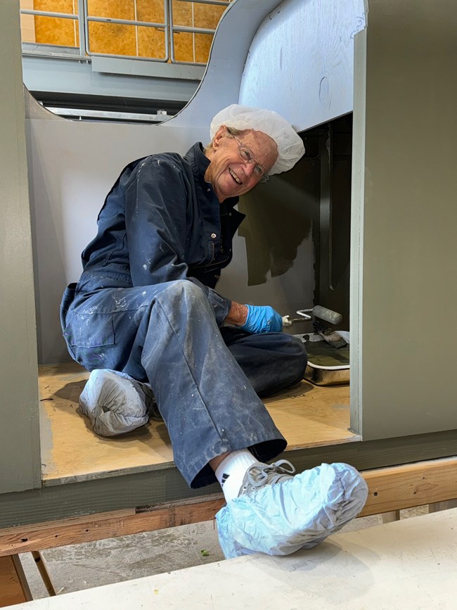 A male volunteer, wearing glasses, coveralls, and paper covers for his hair and shoes, smiles for the camera as he applies olive green paint to wood with a small roller.
