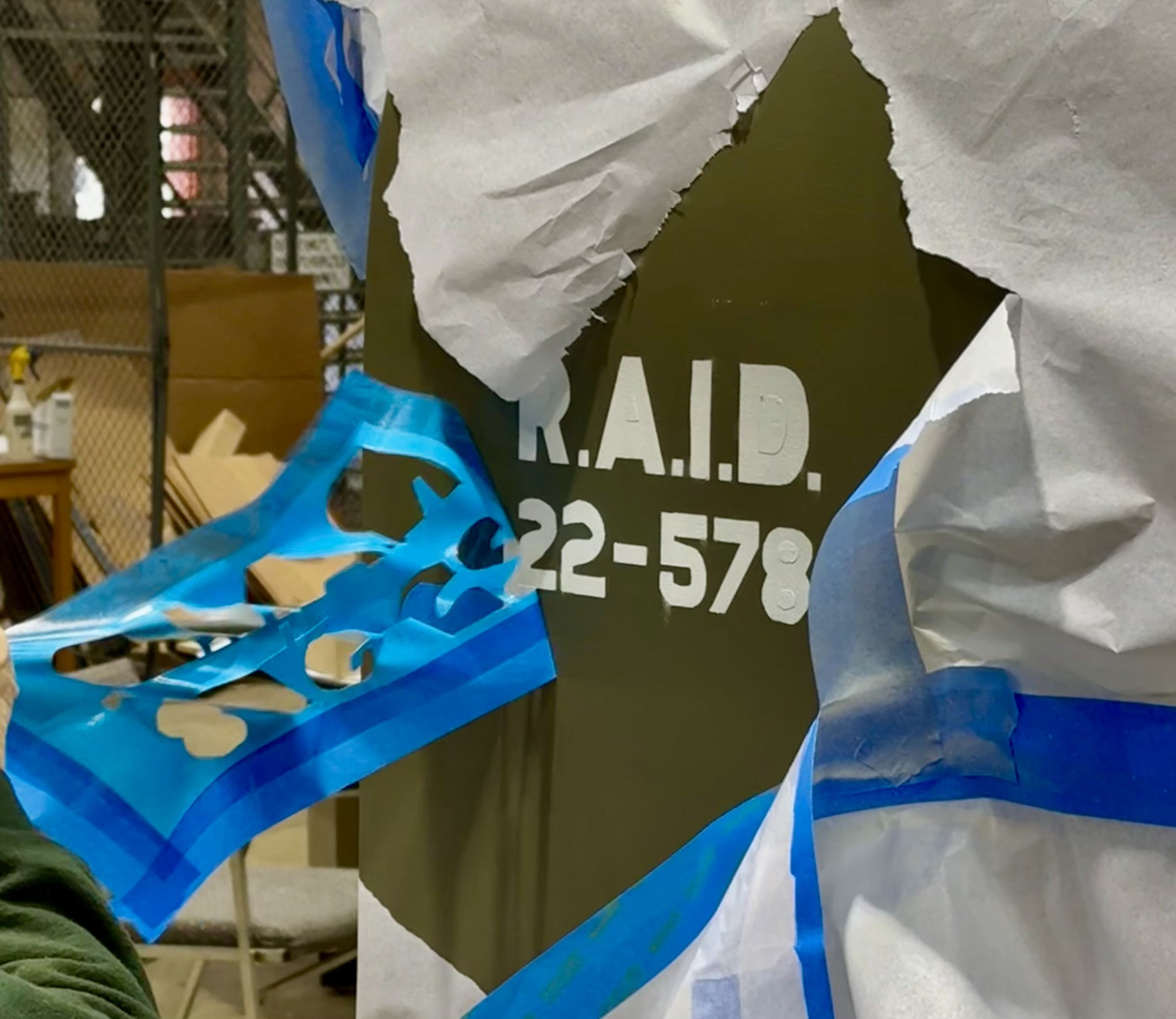 A close-up of a section of the airplane body shows a sticky, blue decal outline being pulled off after painting. White text reading R.A.I.D. 22-578 is on an olive green background.