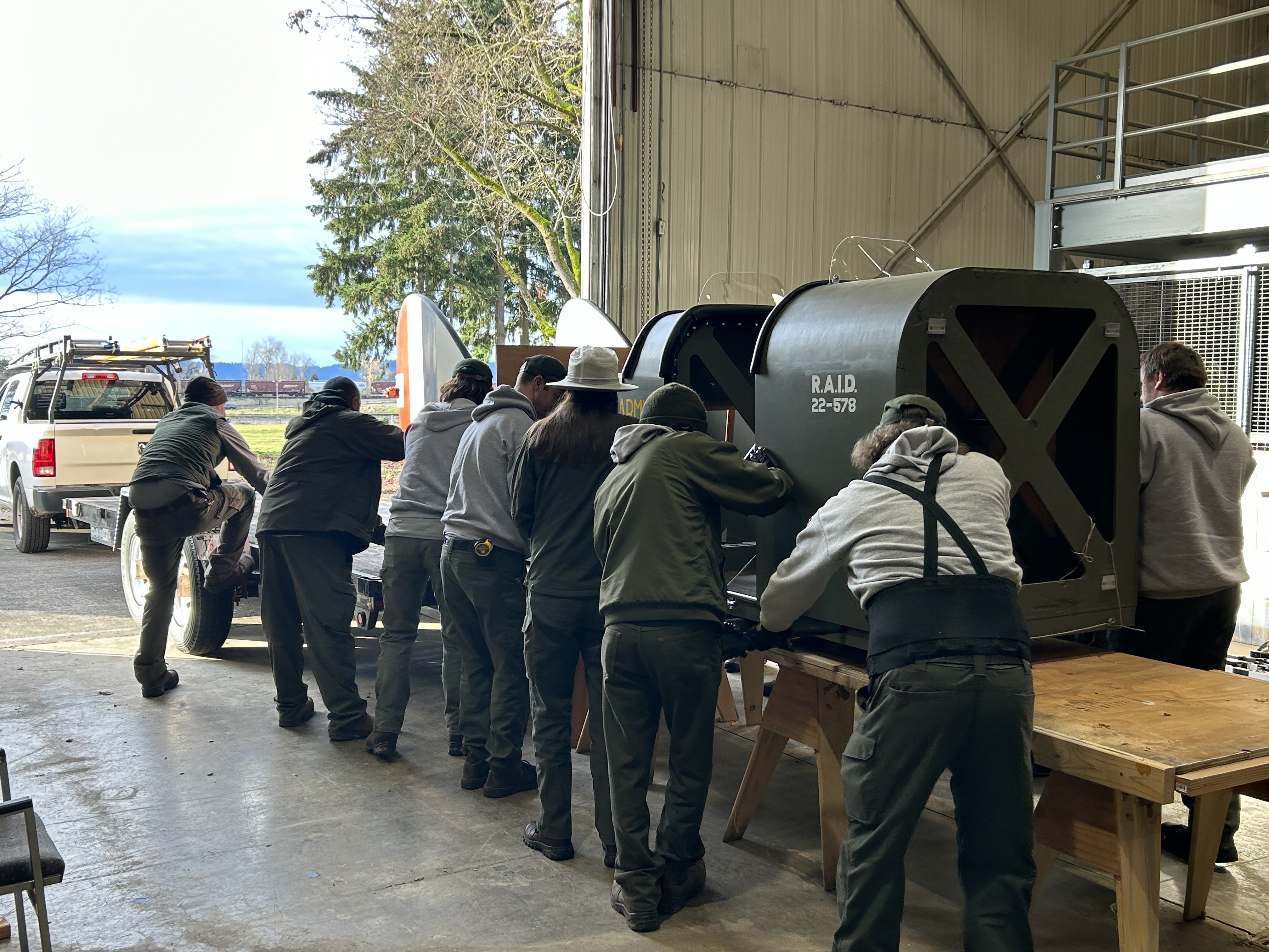 Twelve National Park Service staff, wearing uniforms, lift the aircraft from a wooden support onto a trailer for transport. They are in a warehouse with an open bay door.