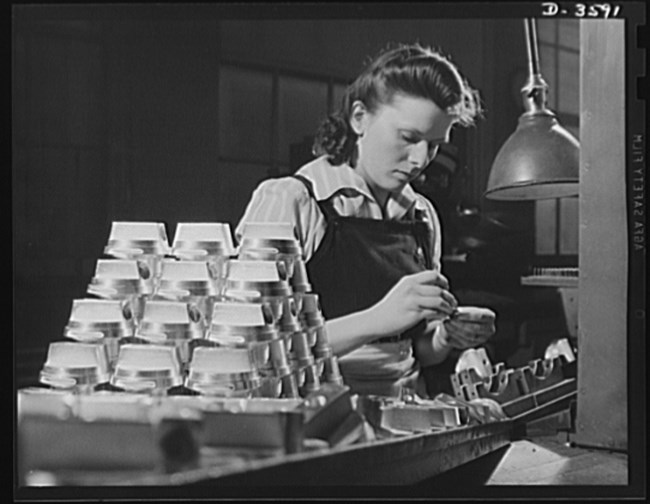 Dark-haired white womans bends her head to assemble part of an ammunition shell. She is wearing an apron and completed shells are stacked in a pyramid to her right