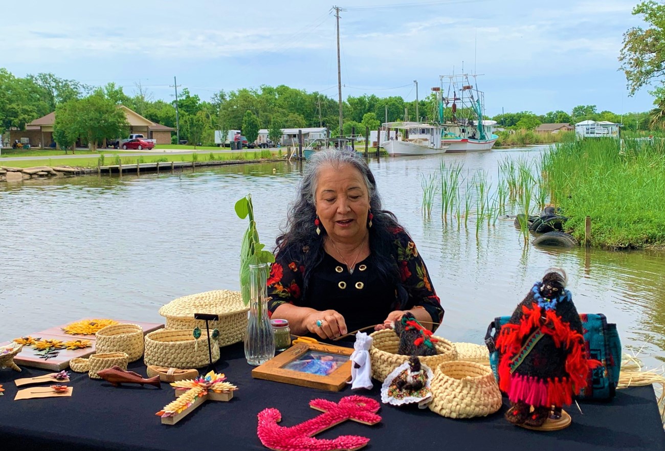 A woman weaving a basket sits at a table in front of a waterway. Table contains various objects representing regional culture including various baskets and dolls.