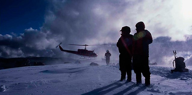 3 people in silhouettes in dusky light in front of a helicopter landing on snowy ground