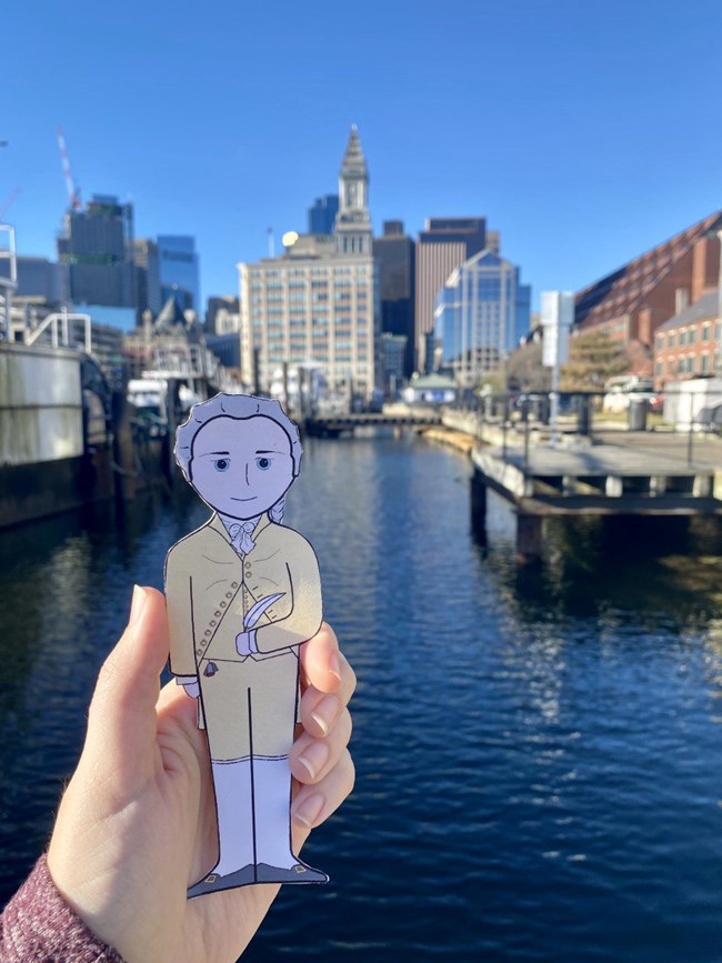 A cartoon illustration cutout of Alexander Hamilton in front of the waters of Boston Harbor, a city scape in the background.