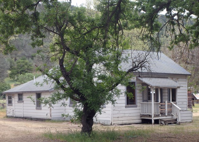 Modest white clapboard, single-story house with small porch and addition on back. Sits in tree-filled mountain valley.