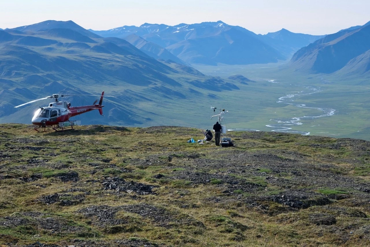 Killik Climate Station in Gates of the Arctic National Park & Preserve A helicopter standby while two people work on a climate station on a bench perched above the Killik valley with mountains and a river in the distance