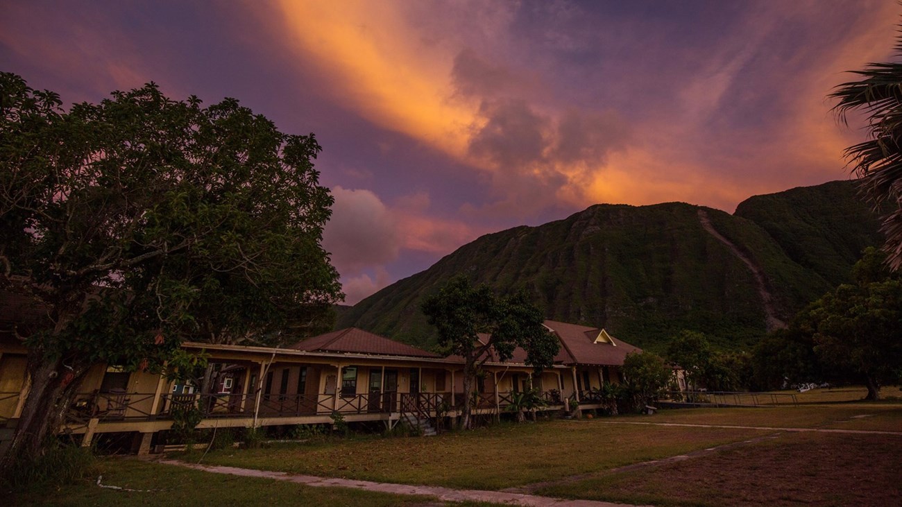 Long, rectangular brown building with continuous porch sits about tropical trees at base of lush mountain at sunset.