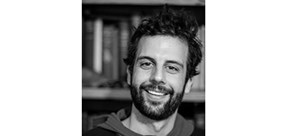 Ben Gaglioti A dark-haired young man smiling at the camera in front of a bookcase of books