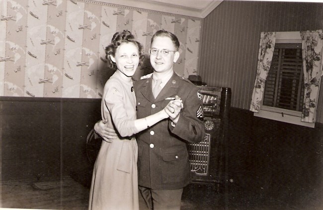 Man in dress uniform and smiling woman dancing in a house.