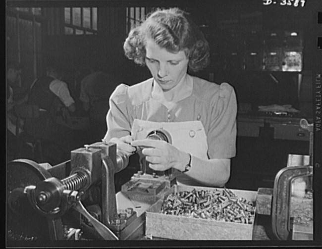 Woman operates a piece of machinery at a worktable. A box of bullet casings is on the table next to her. The woman has curly hair and wears an apron.