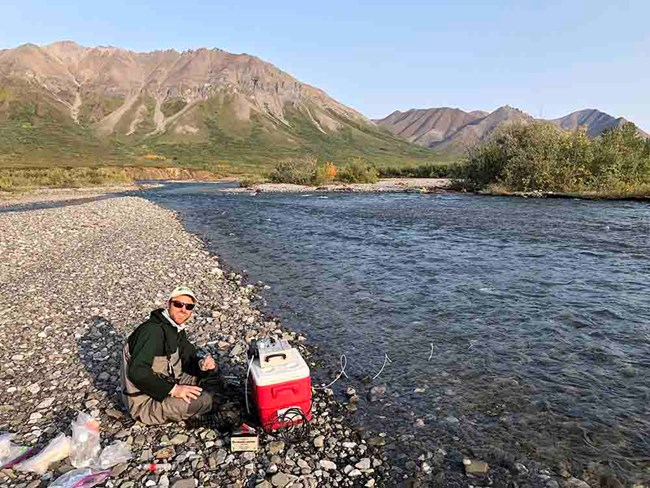 A researcher filters water on a rocky shore along an Arctic river.