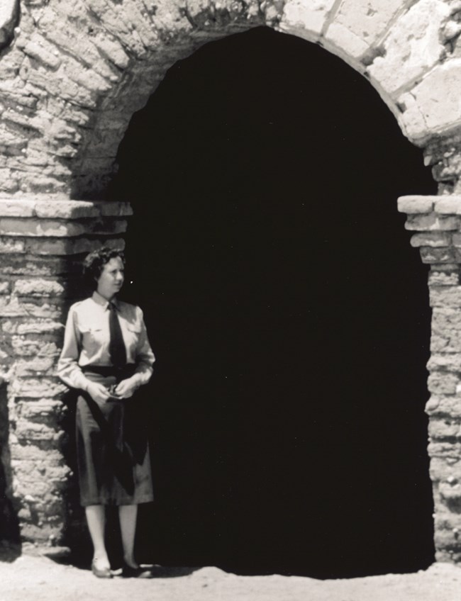 Sallie Brewer, 1947 Sallie Brewer in NPS uniform with skirt stands in front of a stone building.
