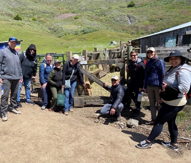 Group of individuals posing for a photo with a goat, a dirt path, and grass in the background.