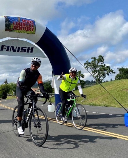 Two males on bicycles. Male on left is wearing dark clothes, while male on right is wearing a bright green neon vest and dark pants. Behind them is a sign that says "finish," grass, and clouds.