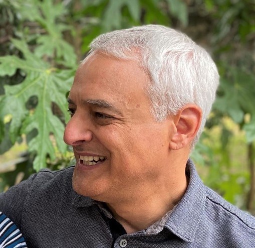 Older Latino male smiling with white hair looking off to the side with a grey shirt and trees in the background.