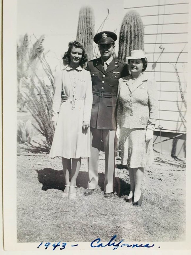 Black and white photo of a white woman in a dress, a white man in military uniform, and an older woman in a skirt set and hat in front of two cacti