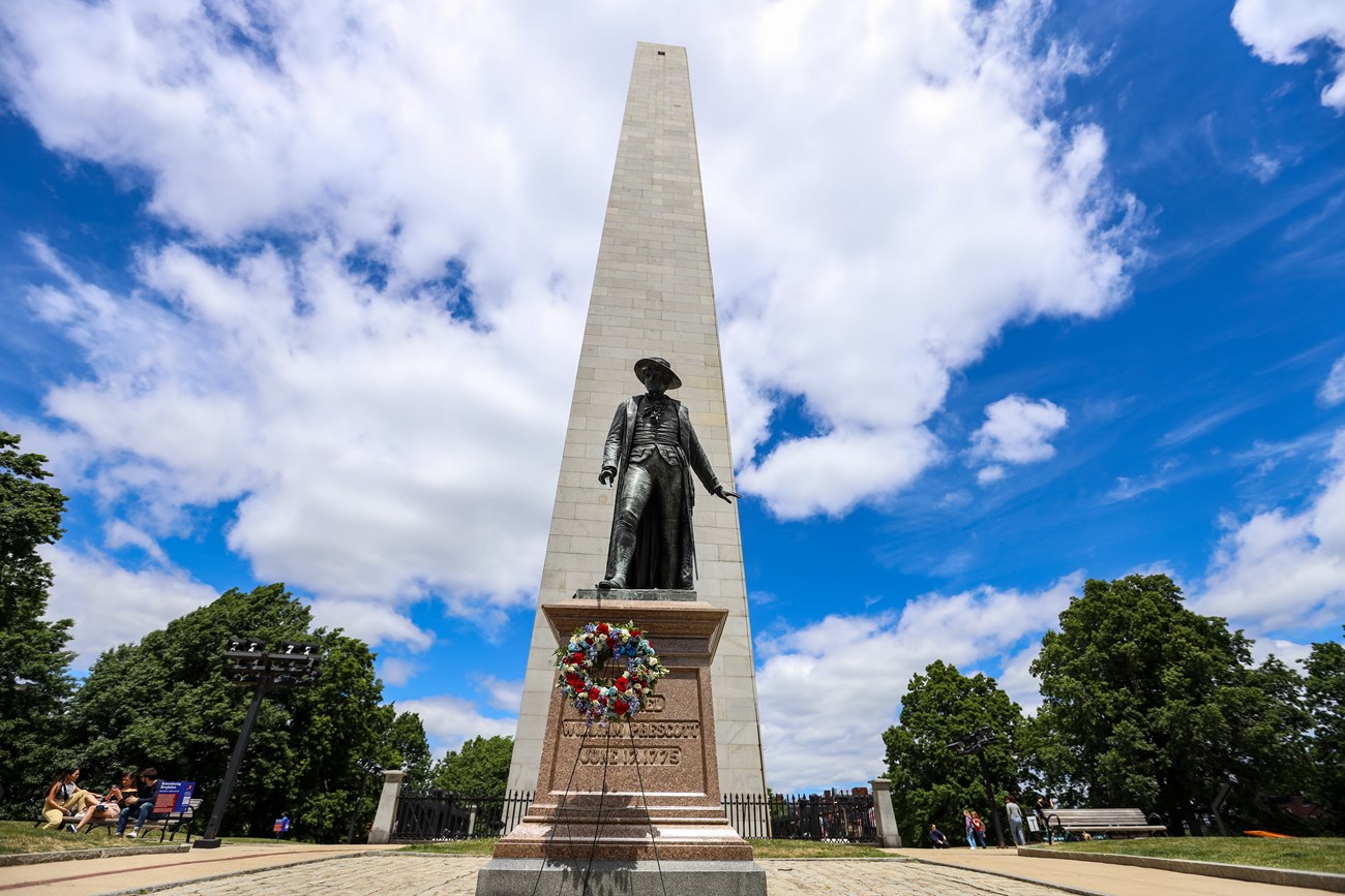 Bunker Hill Monument with the Prescott Statue before it. On a stand in front of the statue is a red, white and blue decorated wreath. A blue sky is filled with white clouds.