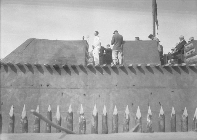 Byrd_at_1927_Fort A man in a crisp white uniform stands in a miniature wooden fort addressing a crowd.