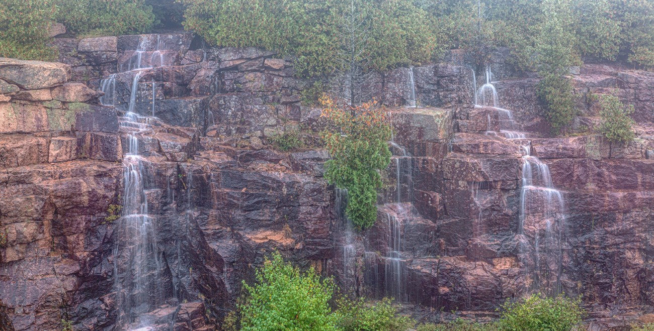 Three separate small waterfalls trickle off a wide fractured cliff punctuated by green shrubbery